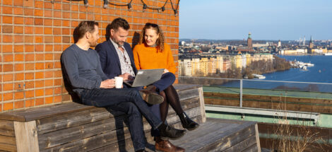 Två killar och en tjej sitter på en terrass med utsikt över Stockholm, och tittar på en dator